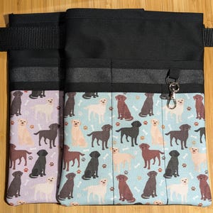 May include: Two black canvas utility belts with multiple pockets. The belt on the left has a purple and white pattern with black labrador retrievers and bones. The belt on the right has a blue and white pattern with black, brown, and yellow labrador retrievers and bones.