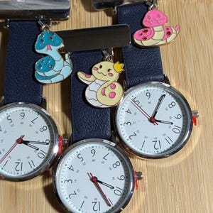 Può includere: Tre orologi da infermiera color argento con quadranti bianchi, numeri neri e lancette dei secondi rosse. Ogni orologio ha un cinturino blu navy e un ciondolo decorativo a forma di serpente blu, giallo o rosa. Su una superficie di legno.