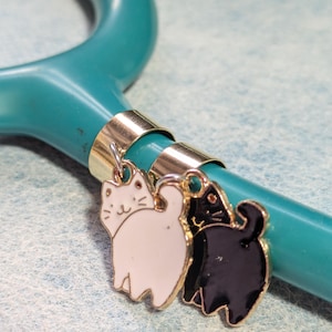 May include: Two gold-toned cat charms attached to a teal stethoscope. The cat charms are facing each other. One cat is white with black eyes and the other is black with white eyes.