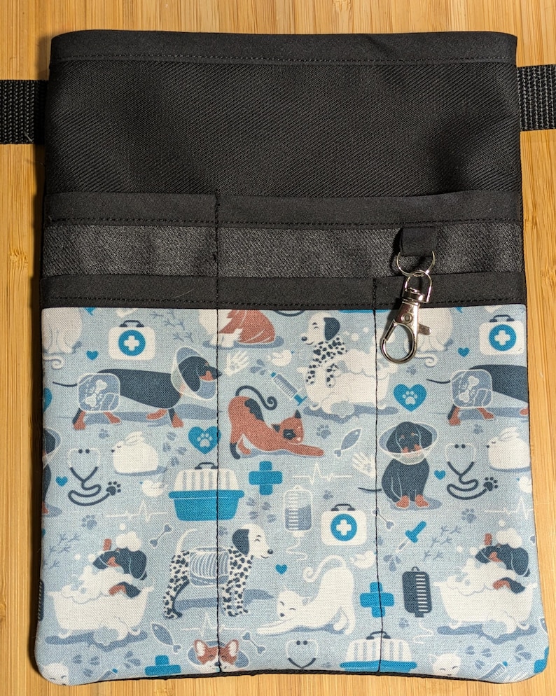May include: Black utility belt with multiple pockets and a clip-on hook. The belt is lined with a blue and white fabric featuring a pattern of dogs, cats, and veterinary symbols.