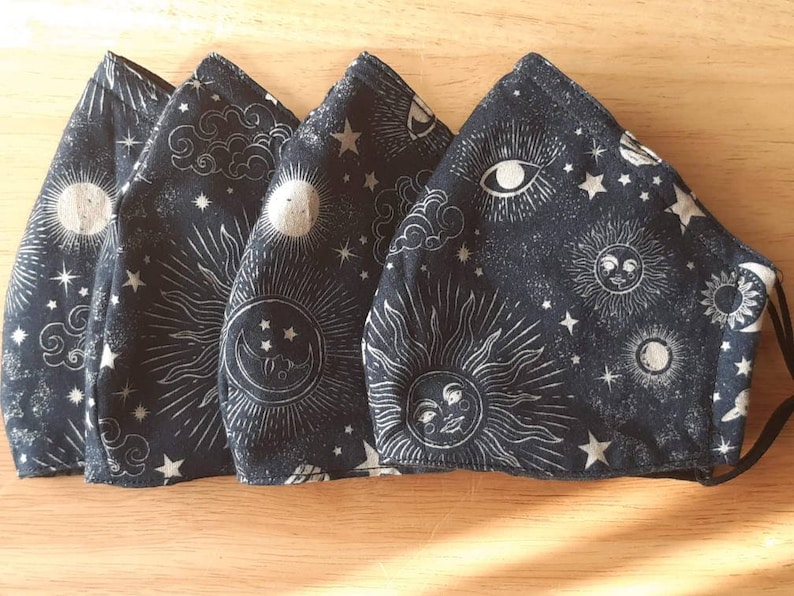 May include: Four black fabric face masks with a white celestial print. The print features suns, moons, stars, and clouds.