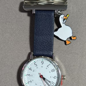 Peut inclure: Une montre infirmière argentée et blanche avec un bracelet en cuir noir et un charme de l'oie. Le cadran de la montre a des aiguilles noires et un fond blanc avec des chiffres noirs et des marques vertes. Le charme de l'oie est blanc avec des accents orange et tient une carotte.