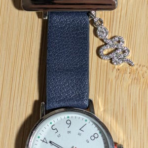 Peut inclure: Une montre de poche d'infirmière argentée avec un bracelet en cuir bleu marine. Le cadran de la montre est blanc avec des chiffres noirs et une trotteuse rouge. Un charme de serpent argenté avec des cristaux transparents est suspendu à la sangle.