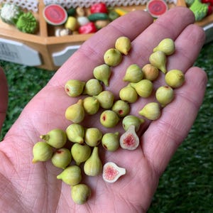 Pu&ograve; includere: Una collezione di fichi in miniatura di colore verde chiaro, che include diversi pezzi interi e due met&agrave; che rivelano un interno testurizzato rosa e bianco. Queste piccole repliche realistiche di cibo sono sparse su un palmo aperto.