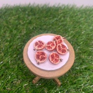 Pu&ograve; includere: Un piattino bianco in miniatura contenente cinque pezzi di melograno tagliati a met&agrave; con semi rossi visibili. Il piatto poggia su un piccolo tavolo rotondo in legno marrone chiaro. Le met&agrave; mostrano un interno strutturato con arilli rossi.