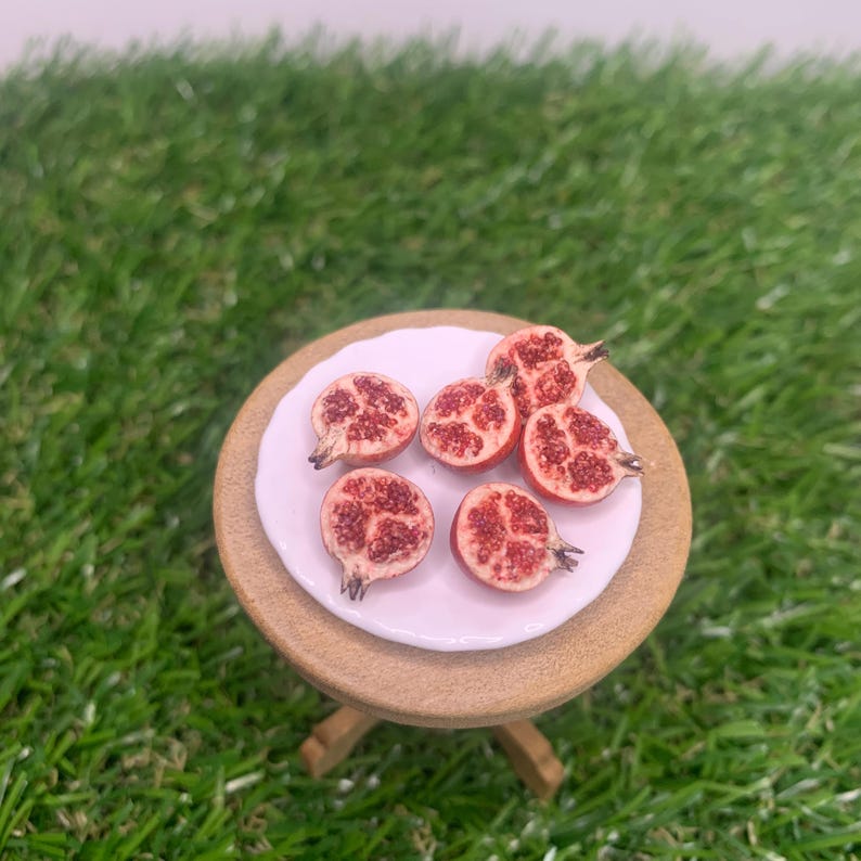 Pu&ograve; includere: Un piattino bianco in miniatura contenente cinque pezzi di melograno tagliati a met&agrave; con semi rossi visibili. Il piatto poggia su un piccolo tavolo rotondo in legno marrone chiaro. Le met&agrave; mostrano un interno strutturato con arilli rossi.