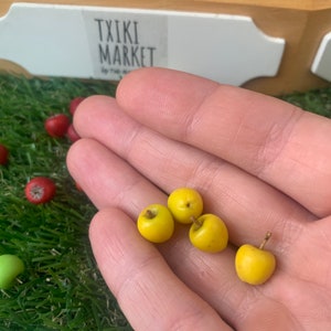 Pu&ograve; includere: Quattro mele gialle in miniatura con piccoli piccioli marroni che riposano in un palmo. Il frutto &egrave; liscio, rotondo e di colore solido. Sullo sfondo, ci sono altre piccole mele rosse e verdi sparse su erba artificiale vicino a un cartello bianco.