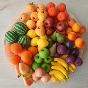 Könnte beinhalten: Eine Sammlung von künstlichen Miniaturfrüchten, die auf einem runden Holzbrett angeordnet sind, darunter kleine Wassermelonen, Orangen, Zitronen, Äpfel, Birnen, Bananen, Pflaumen und Granatäpfel in verschiedenen Farben wie Rot, Gelb, Grün, Orange und Violett.