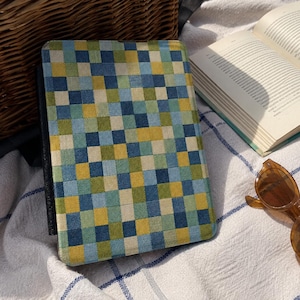 May include: A rectangular electronic tablet case featuring a woven-style grid pattern of small squares in shades of navy blue, light blue, olive green, and yellow. The case has a black spine and a textured fabric surface with a consistent geometric design.