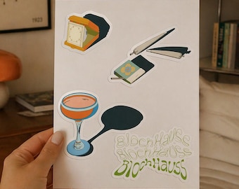 Può includere: Un foglio di adesivi con varie illustrazioni. I disegni includono un bicchiere da cocktail, un orologio, un libro con un fiore e un oggetto arrotolato. Gli adesivi presentano anche il testo "BlochHaus" in un font stilizzato.