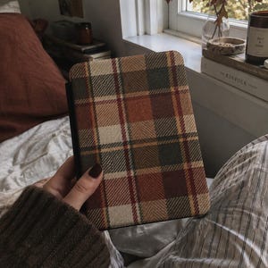 Könnte beinhalten: Ein rechteckiges E-Reader-Gerät in einer Schutzhülle mit gewebter Stoffstruktur und einem mehrfarbigen Karomuster in Braun-, Beige-, Creme- und Dunkelgrüntönen, wobei an der linken Seite ein durchgehend schwarzer Buchrücken zu sehen ist.