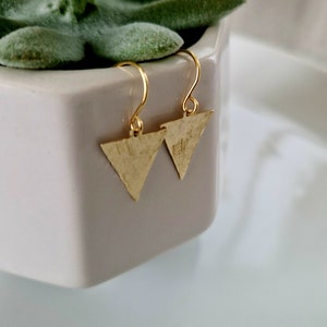 May include: A pair of gold triangle earrings with a hammered texture. The earrings are hanging from a white surface.