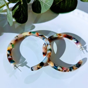 Puede incluir: Un par de pendientes grandes de aro hechos de plástico marmoleado de colores. Los pendientes son de color marrón claro con salpicaduras de azul, verde y rosa.