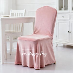 Puede incluir: Una funda de silla de terciopelo rosa con respaldo redondeado y parte inferior con volantes. El texto "For Happy Day" está escrito en cursiva en la parte delantera. La funda está sobre una silla de madera.