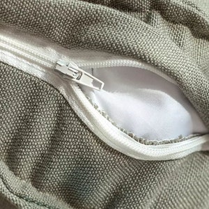 May include: Close-up of a zipper on a grey fabric cushion cover. The zipper is white and has a metal slider.