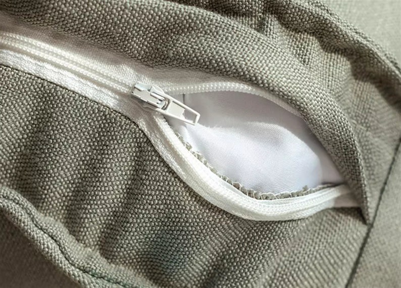 May include: Close-up of a zipper on a grey fabric cushion cover. The zipper is white and has a metal slider.