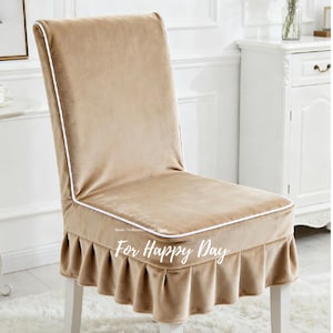 May include: Beige chair cover with white piping and a ruffled skirt. The cover is made of a soft, velvety material and is designed to fit over a standard chair. The text "For Happy Day" is printed on the skirt.