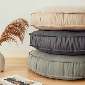 May include: Three round floor cushions stacked on top of each other. The bottom cushion is a light green colour, the middle cushion is a dark grey colour, and the top cushion is a light beige colour. The cushions are made of a soft, textured fabric.