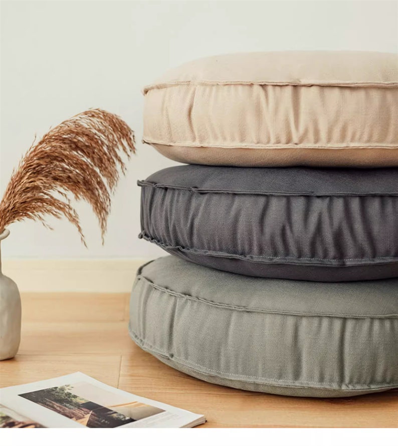 May include: Three round floor cushions stacked on top of each other. The bottom cushion is a light green colour, the middle cushion is a dark grey colour, and the top cushion is a light beige colour. The cushions are made of a soft, textured fabric.