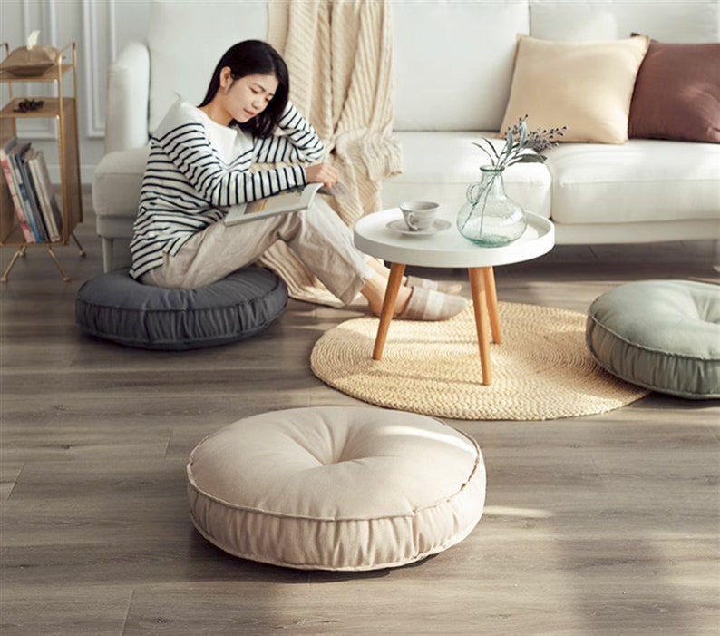 May include: A woman sits on a grey round floor cushion in a living room with a white sofa, a coffee table with a vase of greenery, and two other round floor cushions, one grey and one beige.