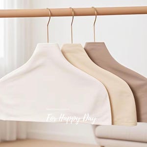 May include: Three padded clothing hangers in off-white, beige, and taupe hang from a wooden rod. The hangers have gold-colored hooks and are designed to protect garments. The phrase "For Happy Day" is printed on the front of the off-white hanger.