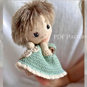 May include: A handmade crochet doll with light brown hair, dark eyes, and a beige complexion. The doll wears a light mint green dress with a cream-colored trim and a small bow detail. The text PDF PATTERN is visible on the right side.