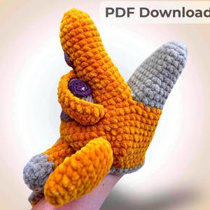 May include: A crochet hand puppet featuring a bright orange body with grey accents on the tips and purple circular patches. The puppet has a textured, soft yarn appearance and includes a rectangular label in the top right corner with the text PDF Download.