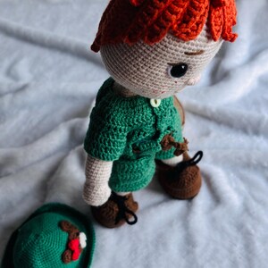 May include: A handmade crochet doll with bright orange hair, wearing a green shirt and brown shoes with black laces. The doll has a light beige complexion and dark eyes. A separate green hat with a small red and white decorative accent sits nearby.