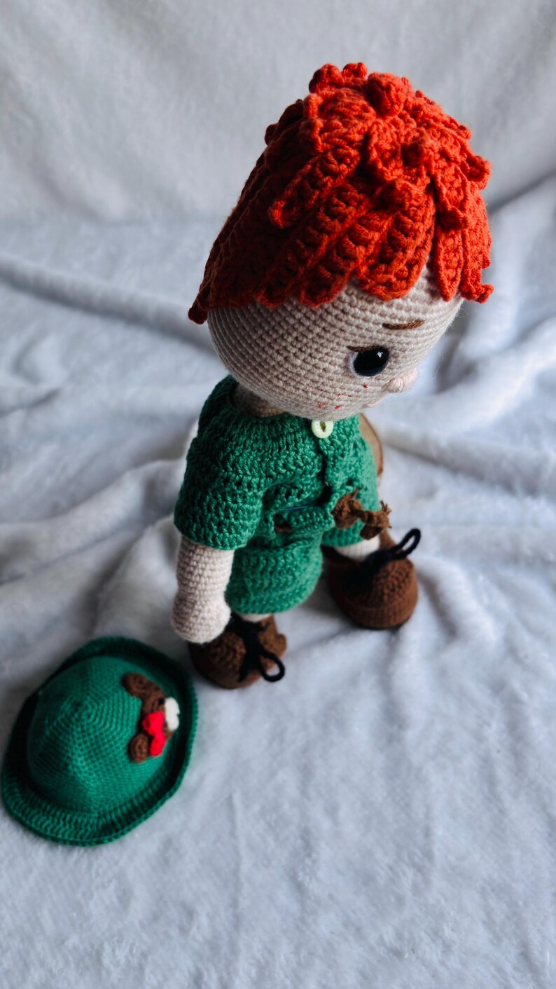 May include: A handmade crochet doll with bright orange hair, wearing a green shirt and brown shoes with black laces. The doll has a light beige complexion and dark eyes. A separate green hat with a small red and white decorative accent sits nearby.