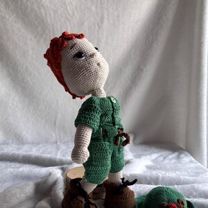 May include: A handmade crochet doll with light skin, textured red hair, and large dark eyes. The doll wears a green short-sleeved jumpsuit with a small button at the neck and brown shoes with black laces. A separate green hat with a red accent sits nearby.
