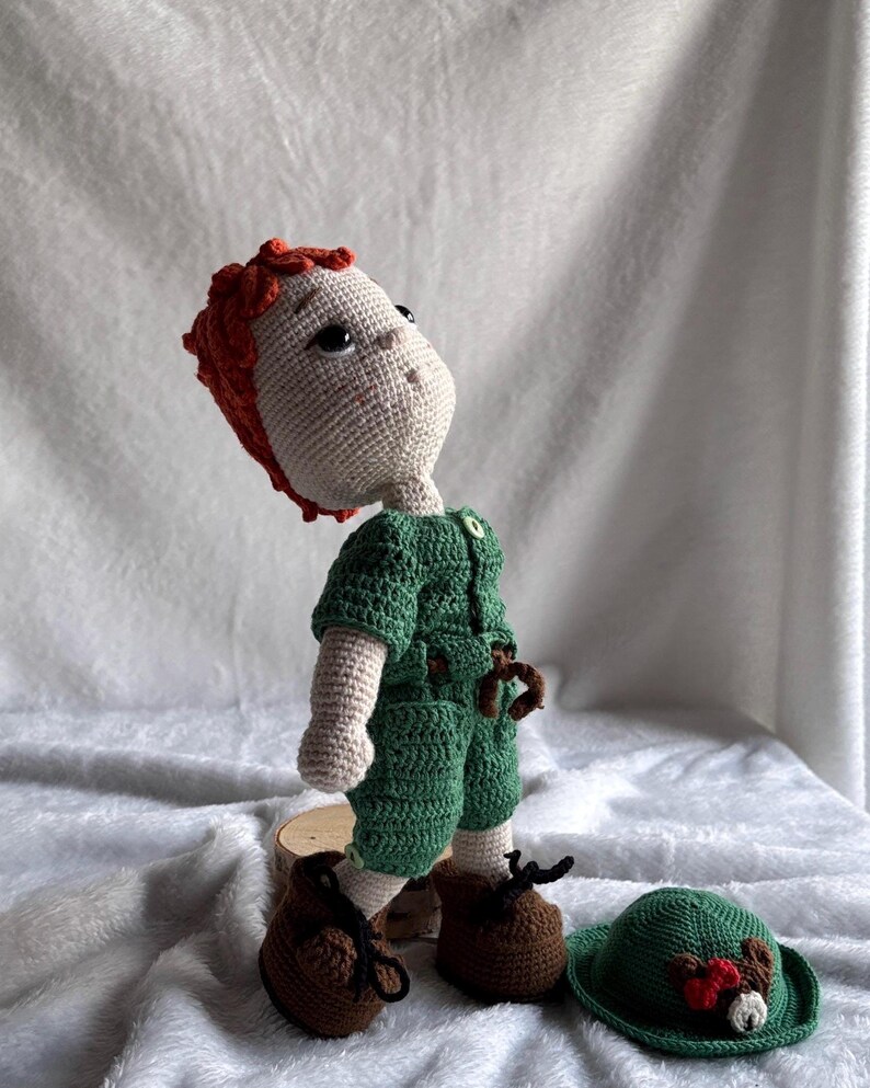 May include: A handmade crochet doll with light skin, textured red hair, and large dark eyes. The doll wears a green short-sleeved jumpsuit with a small button at the neck and brown shoes with black laces. A separate green hat with a red accent sits nearby.