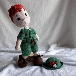 May include: A handmade crochet doll with bright red hair, a light beige complexion, and dark eyes. The doll wears a green short-sleeved shirt with a small button and matching green shorts. It has brown shoes with black laces and a separate green hat.