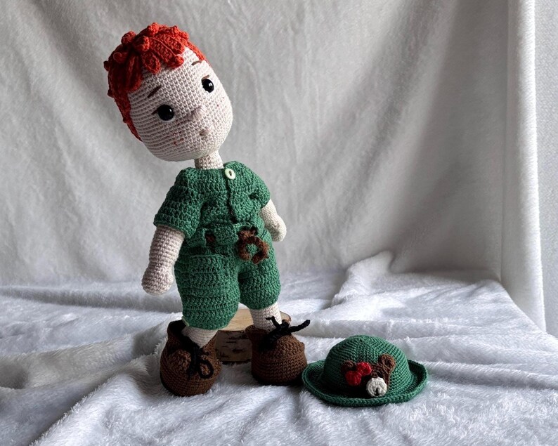May include: A handmade crochet doll with bright red hair, a light beige complexion, and dark eyes. The doll wears a green short-sleeved shirt with a small button and matching green shorts. It has brown shoes with black laces and a separate green hat.