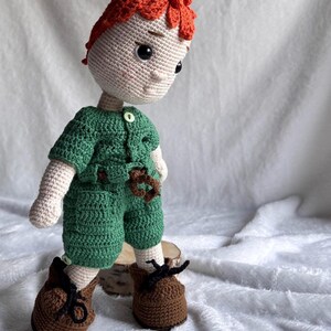 May include: A handmade crochet doll featuring bright orange hair, a light beige complexion, and dark eyes. The doll wears a green short-sleeved shirt with a white button, matching green shorts with a brown belt, and brown shoes with black laces.