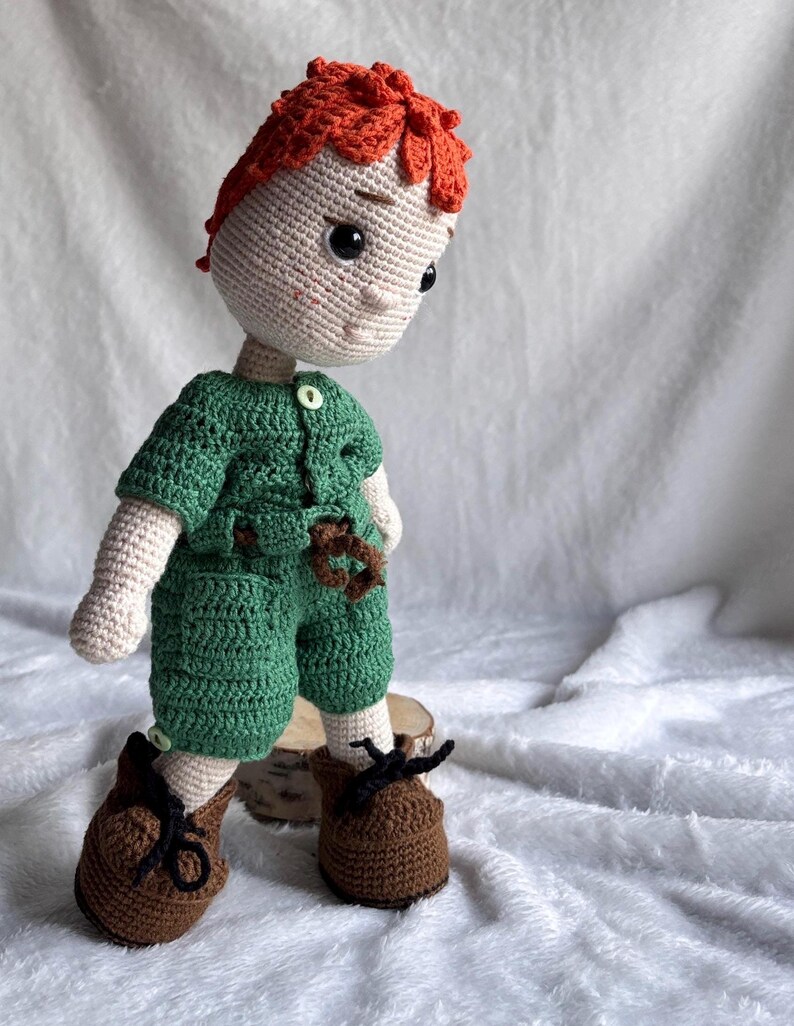 May include: A handmade crochet doll featuring bright orange hair, a light beige complexion, and dark eyes. The doll wears a green short-sleeved shirt with a white button, matching green shorts with a brown belt, and brown shoes with black laces.
