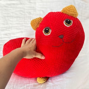 Può includere: Un peluche lavorato all'uncinetto di colore rosso brillante a forma di gatto con due orecchie giallo senape. Il gatto presenta grandi occhi rotondi con pupille marrone scuro, un piccolo naso ricamato in marrone e una bocca semplice.