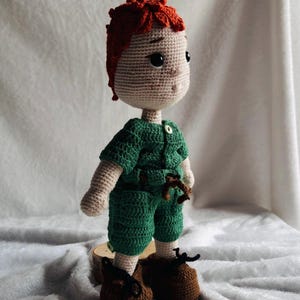 May include: A handmade crochet doll featuring a light beige complexion, textured orange hair, and dark eyes. The doll wears a green short-sleeved romper with a small button at the collar and a brown tie at the waist, paired with brown shoes with black laces.