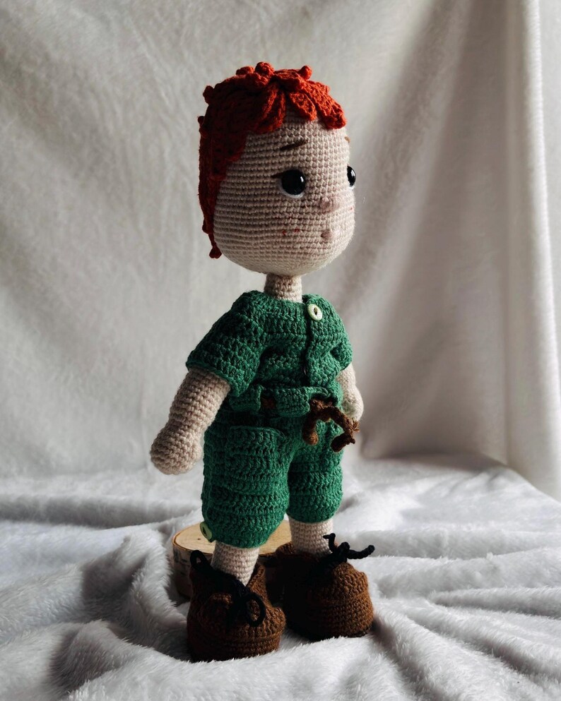 May include: A handmade crochet doll featuring a light beige complexion, textured orange hair, and dark eyes. The doll wears a green short-sleeved romper with a small button at the collar and a brown tie at the waist, paired with brown shoes with black laces.