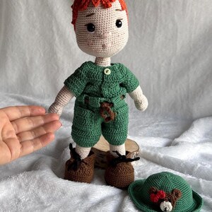 May include: A handmade crochet doll with orange hair, wearing a green short-sleeved shirt and shorts, brown shoes, and a small brown bear accessory on the front. A matching green hat with a bear face and red bow detail is placed next to the doll.