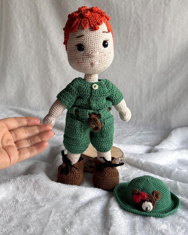 May include: A handmade crochet doll with orange hair, wearing a green short-sleeved shirt and shorts, brown shoes, and a small brown bear accessory on the front. A matching green hat with a bear face and red bow detail is placed next to the doll.