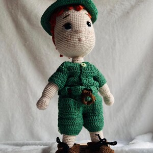 May include: A handmade crochet doll featuring a light beige complexion, orange hair, and a green hat. The doll wears a green short-sleeved shirt with a small button, matching green shorts with a brown belt detail, and brown shoes with black laces.