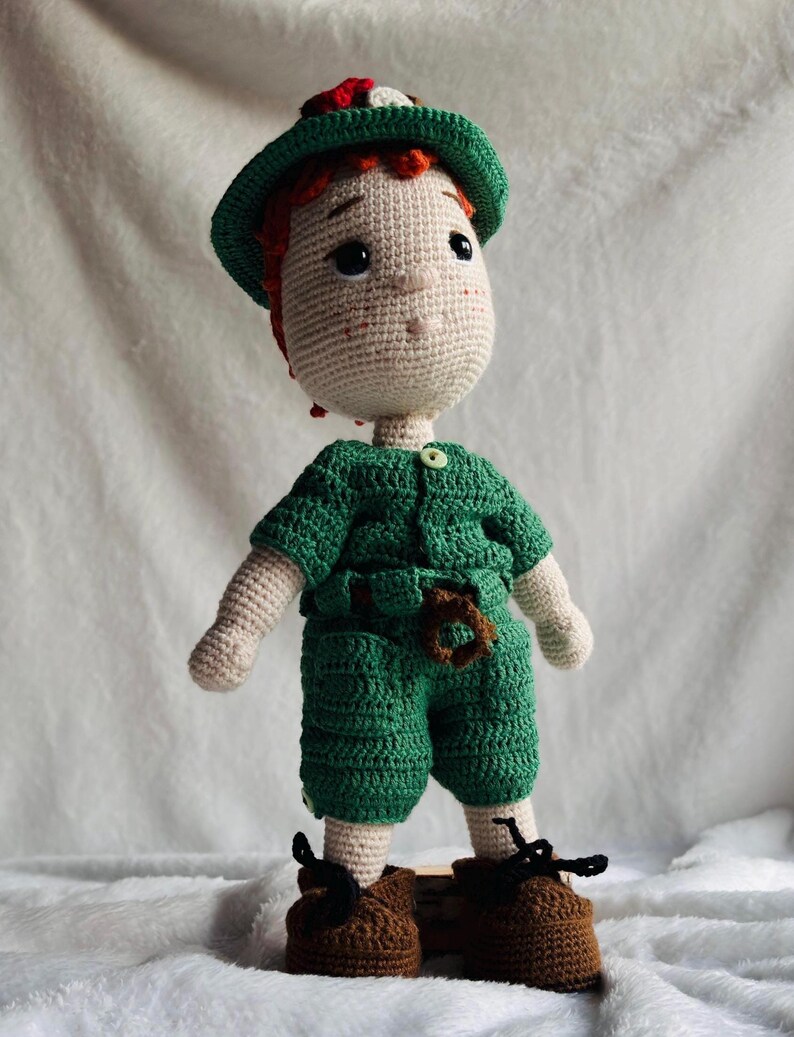 May include: A handmade crochet doll featuring a light beige complexion, orange hair, and a green hat. The doll wears a green short-sleeved shirt with a small button, matching green shorts with a brown belt detail, and brown shoes with black laces.