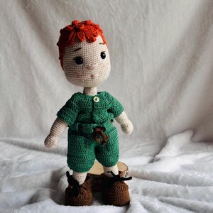 May include: A handmade crochet doll featuring bright orange hair, a light beige complexion, and large black eyes. The doll wears a green short-sleeved shirt with a small button, matching green shorts with a brown belt detail, and brown shoes.