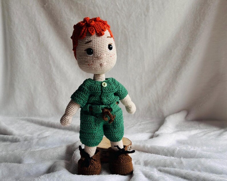 May include: A handmade crochet doll featuring bright orange hair, a light beige complexion, and large black eyes. The doll wears a green short-sleeved shirt with a small button, matching green shorts with a brown belt detail, and brown shoes.