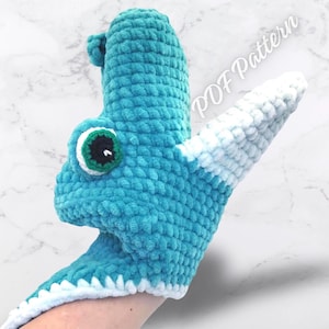 May include: A handmade, turquoise crochet dragon puppet with a white and light blue striped tail and a large, round eye with a black pupil. The puppet has a small horn and the text "PDF Pattern" is visible.