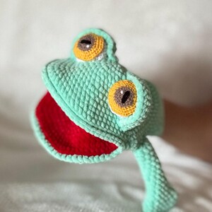 May include: A crochet frog hand puppet featuring a light green textured body, a bright red open mouth, and two prominent eyes with yellow irises and dark brown pupils. The puppet is crafted with visible yarn stitches throughout its entire surface.