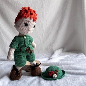 May include: A handmade crochet doll with bright orange hair, wearing a green short-sleeved shirt and matching shorts with a small white button. The doll has brown shoes with black laces and stands next to a small green hat featuring a bear face.