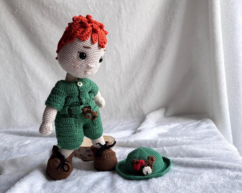 May include: A handmade crochet doll with bright orange hair, wearing a green short-sleeved shirt and matching shorts with a small white button. The doll has brown shoes with black laces and stands next to a small green hat featuring a bear face.