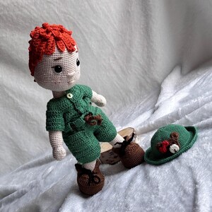 May include: A handmade crochet doll with bright orange hair, wearing a green short-sleeved shirt and matching shorts. The doll has brown shoes with black laces. A separate green hat with a small brown bear applique and red bow is placed next to the doll.
