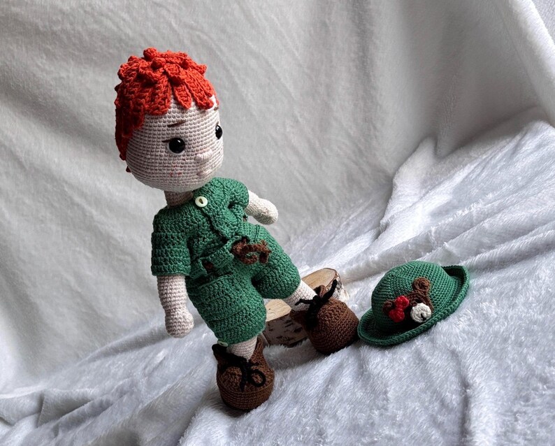 May include: A handmade crochet doll with bright orange hair, wearing a green short-sleeved shirt and matching shorts. The doll has brown shoes with black laces. A separate green hat with a small brown bear applique and red bow is placed next to the doll.